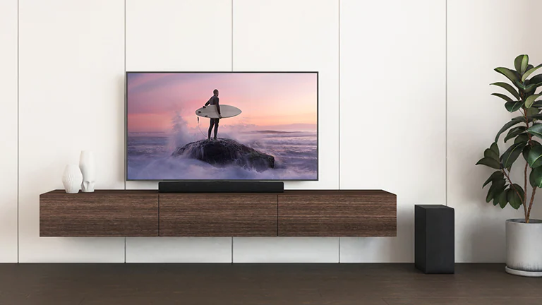 Телевизор и саундбар LG размещены на коричневой полке, а сабвуфер&nbsp;&mdash; на полу. На экране телевизора изображен серфер на скале.